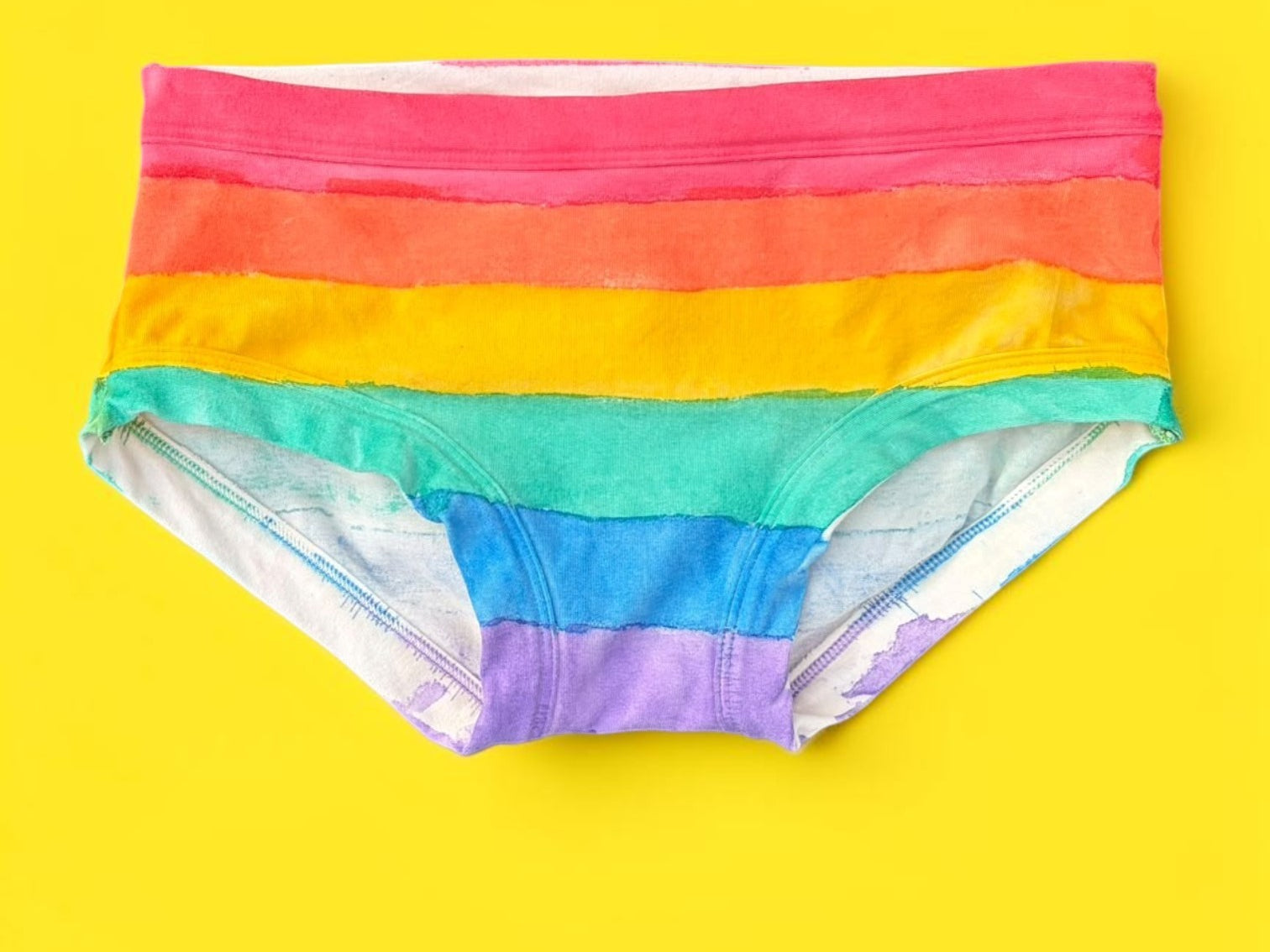 A pair of women's low-rise pants featuring horizontal rainbow stripes in red, orange, yellow, green, blue, and purple, laid flat on a solid bright yellow background.