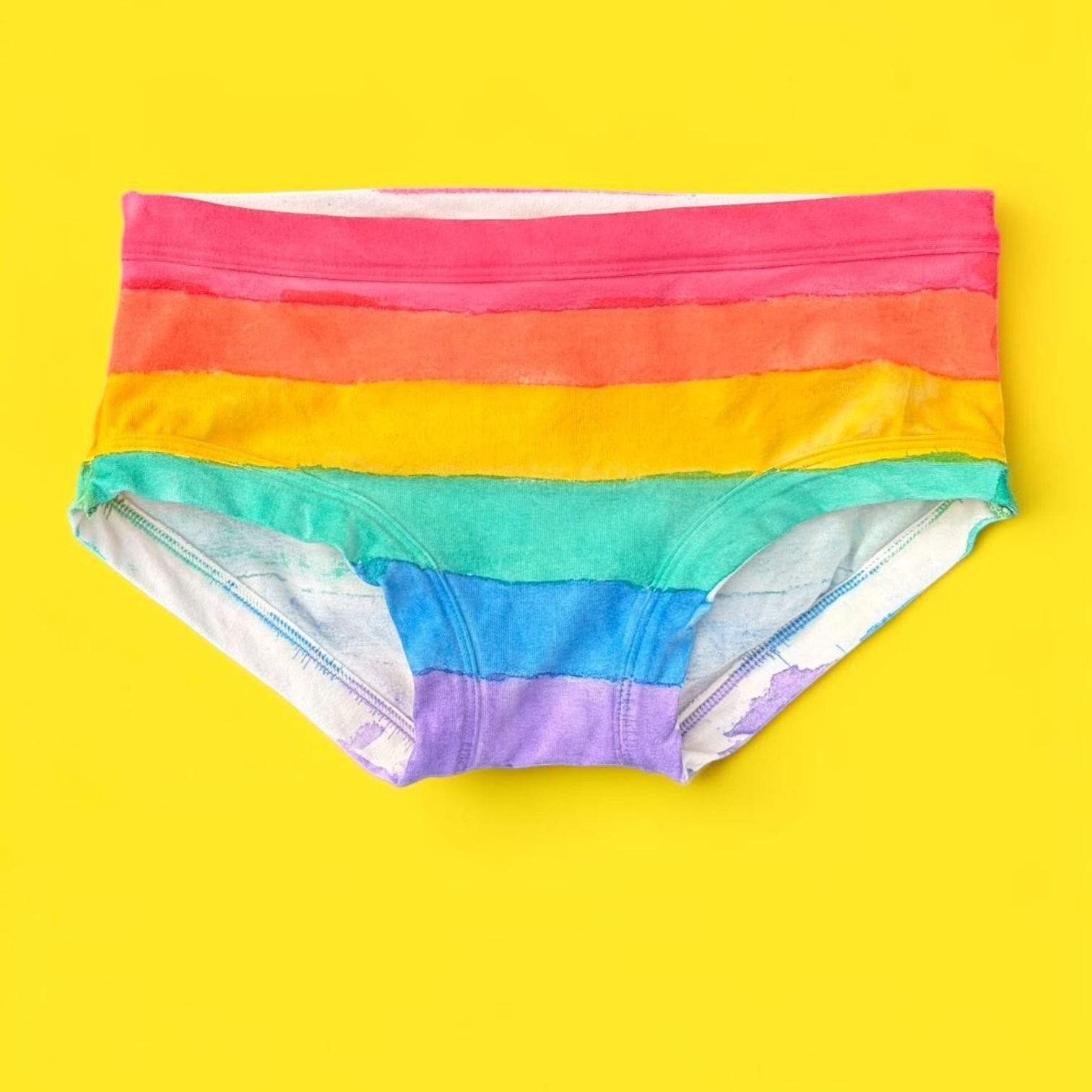 A pair of women's low-rise pants featuring horizontal rainbow stripes in red, orange, yellow, green, blue, and purple, laid flat on a solid bright yellow background.