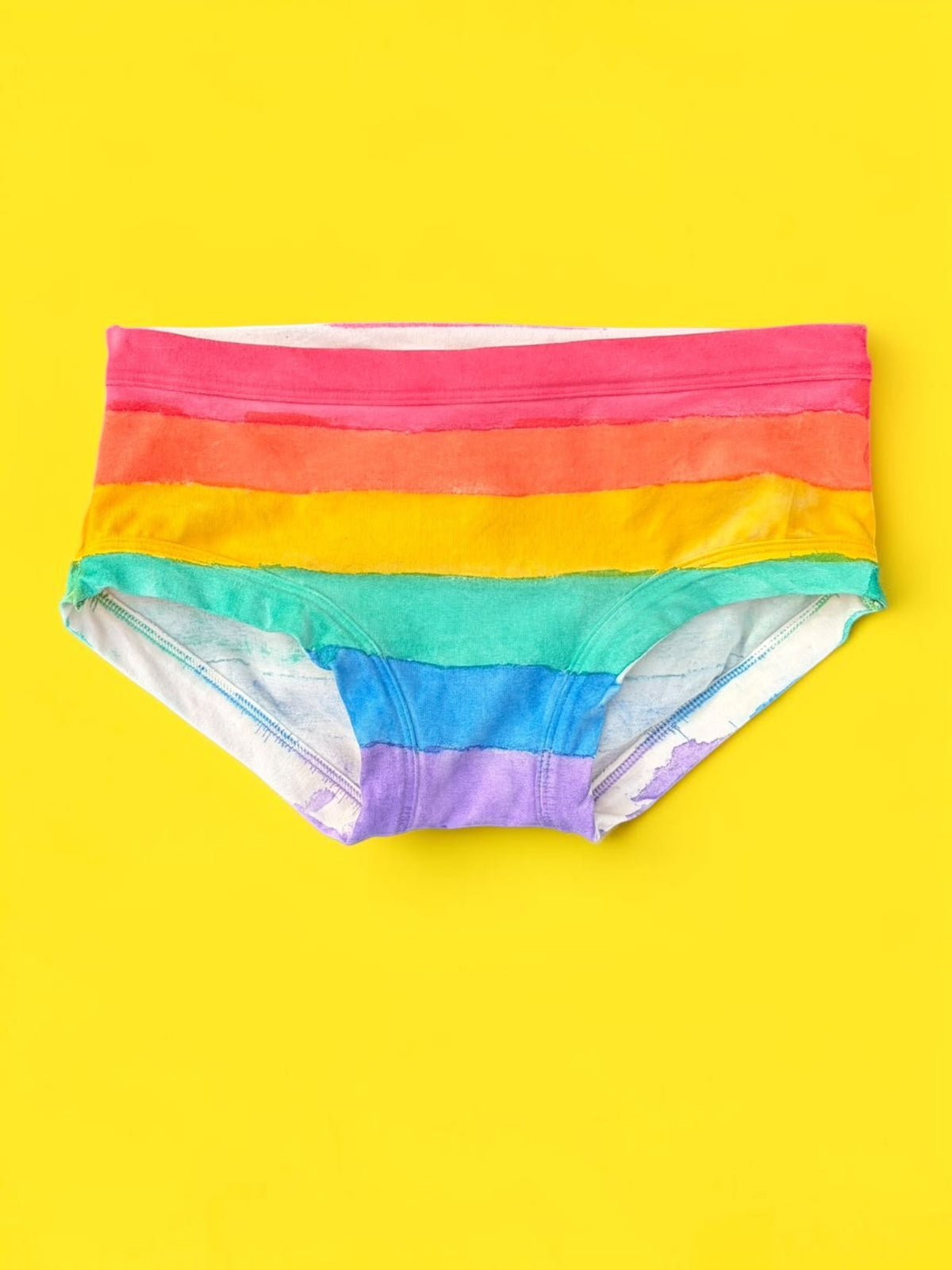 A pair of women's low-rise pants featuring horizontal rainbow stripes in red, orange, yellow, green, blue, and purple, laid flat on a solid bright yellow background.