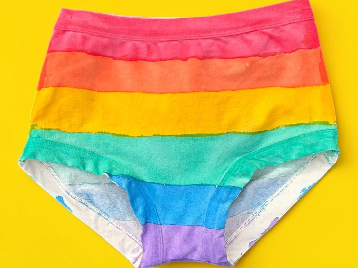 Rainbow-striped hand painted originals laid flat on a bright yellow background. The underwear features horizontal bands of red, orange, yellow, green, blue, and purple, with a white lining visible on the inner legs.