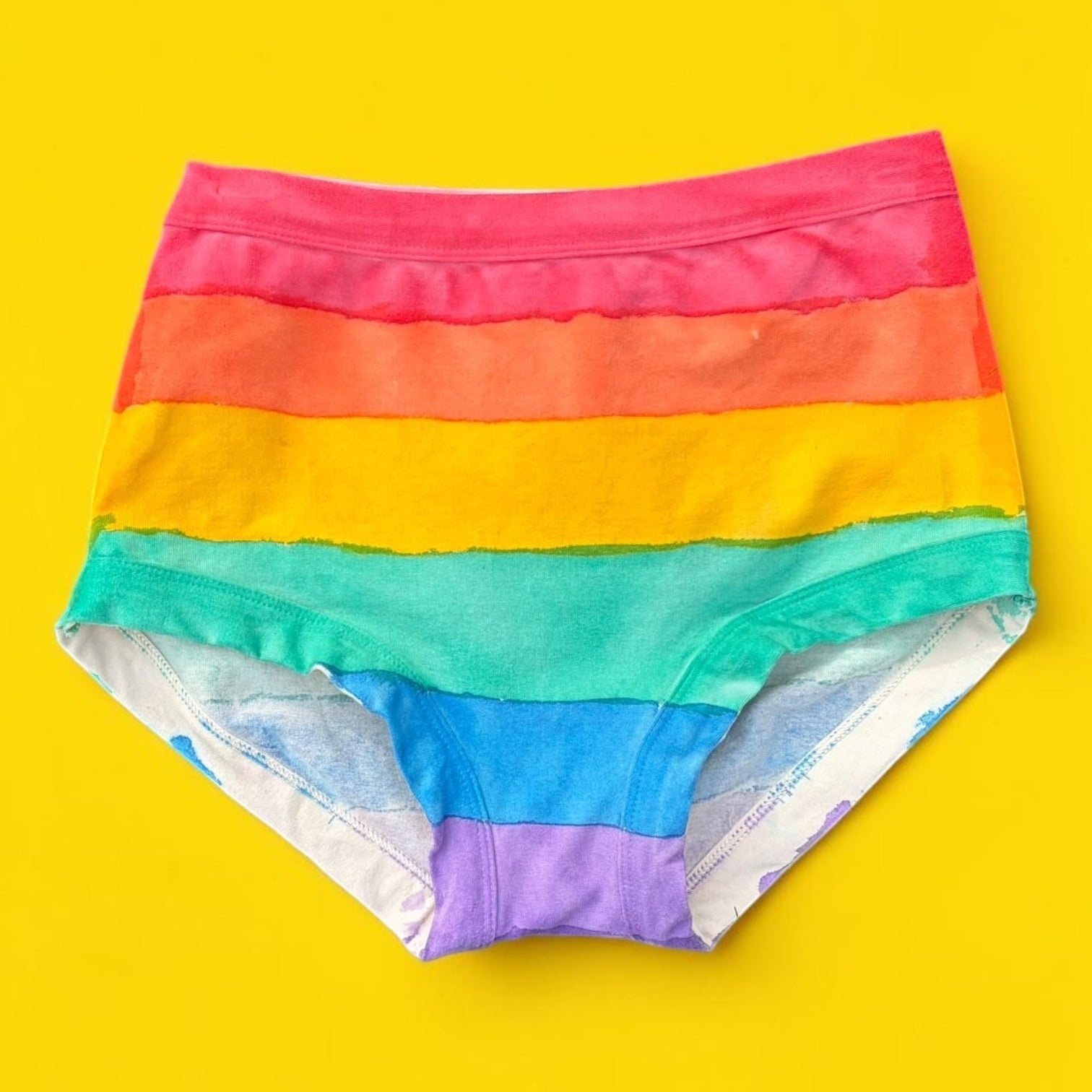 Rainbow-striped hand painted originals laid flat on a bright yellow background. The underwear features horizontal bands of red, orange, yellow, green, blue, and purple, with a white lining visible on the inner legs.
