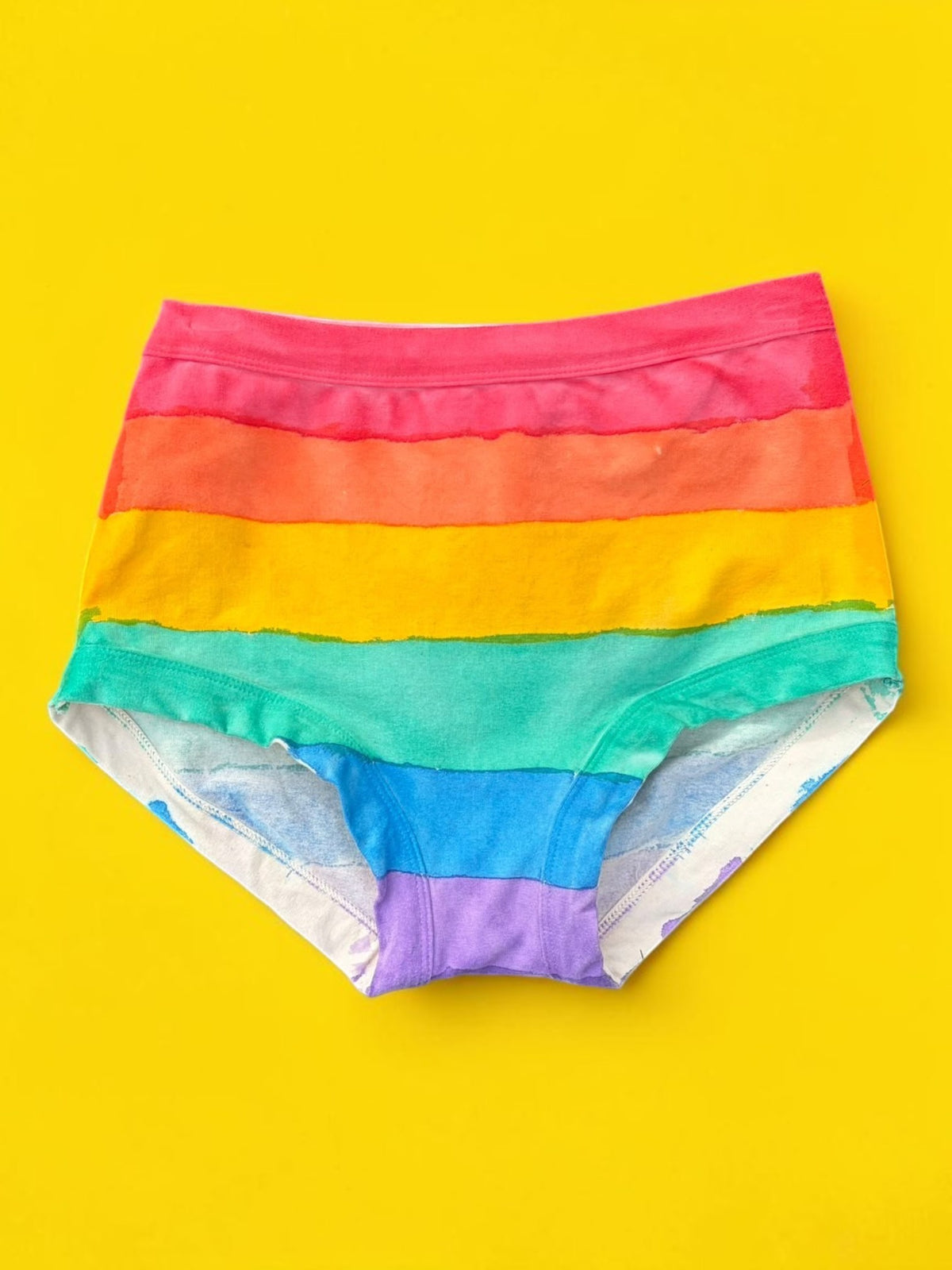 Rainbow-striped hand painted originals laid flat on a bright yellow background. The underwear features horizontal bands of red, orange, yellow, green, blue, and purple, with a white lining visible on the inner legs.