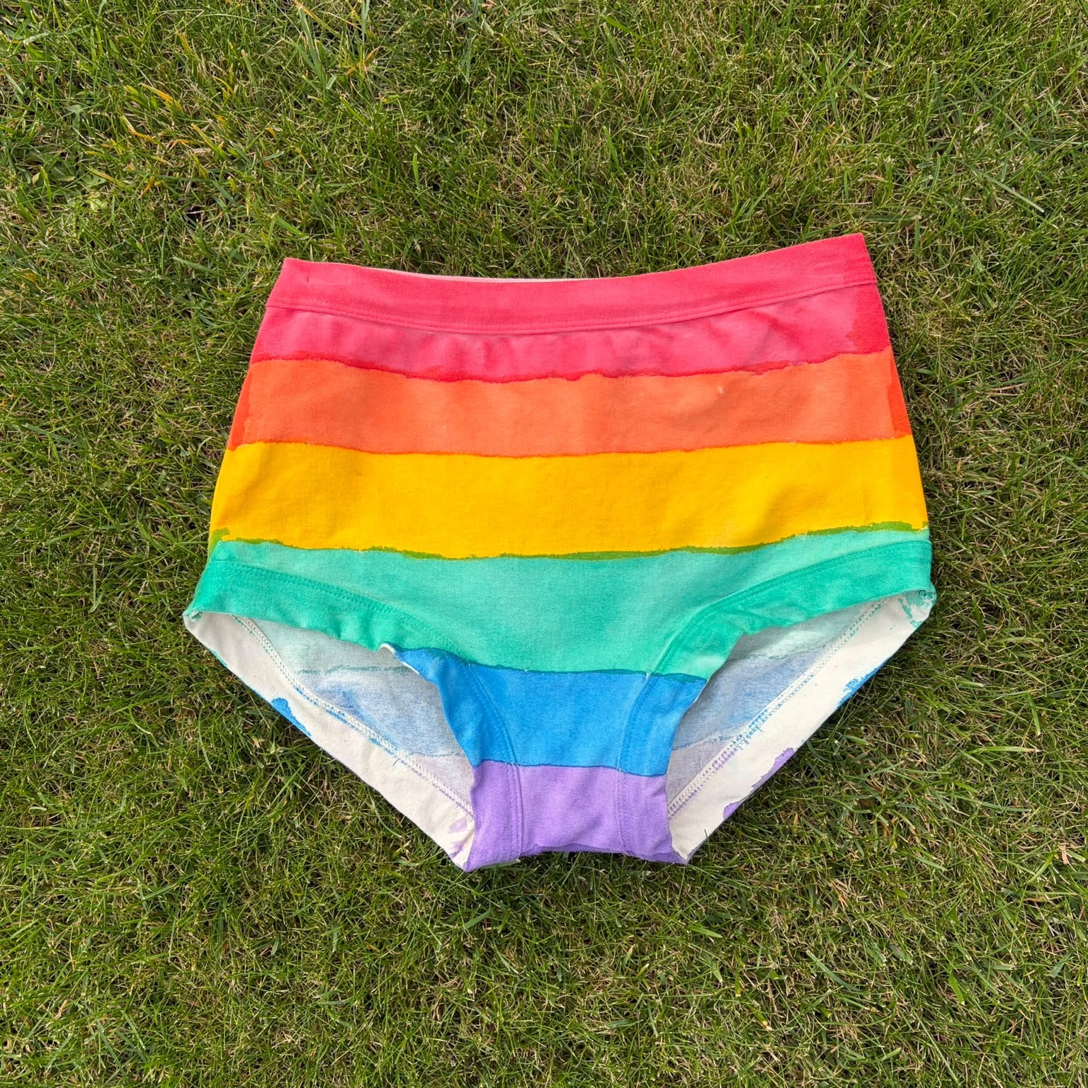 Rainbow-striped original Thunderpants laid flat on green grass, featuring horizontal bands of pink, orange, yellow, green, blue, and purple.