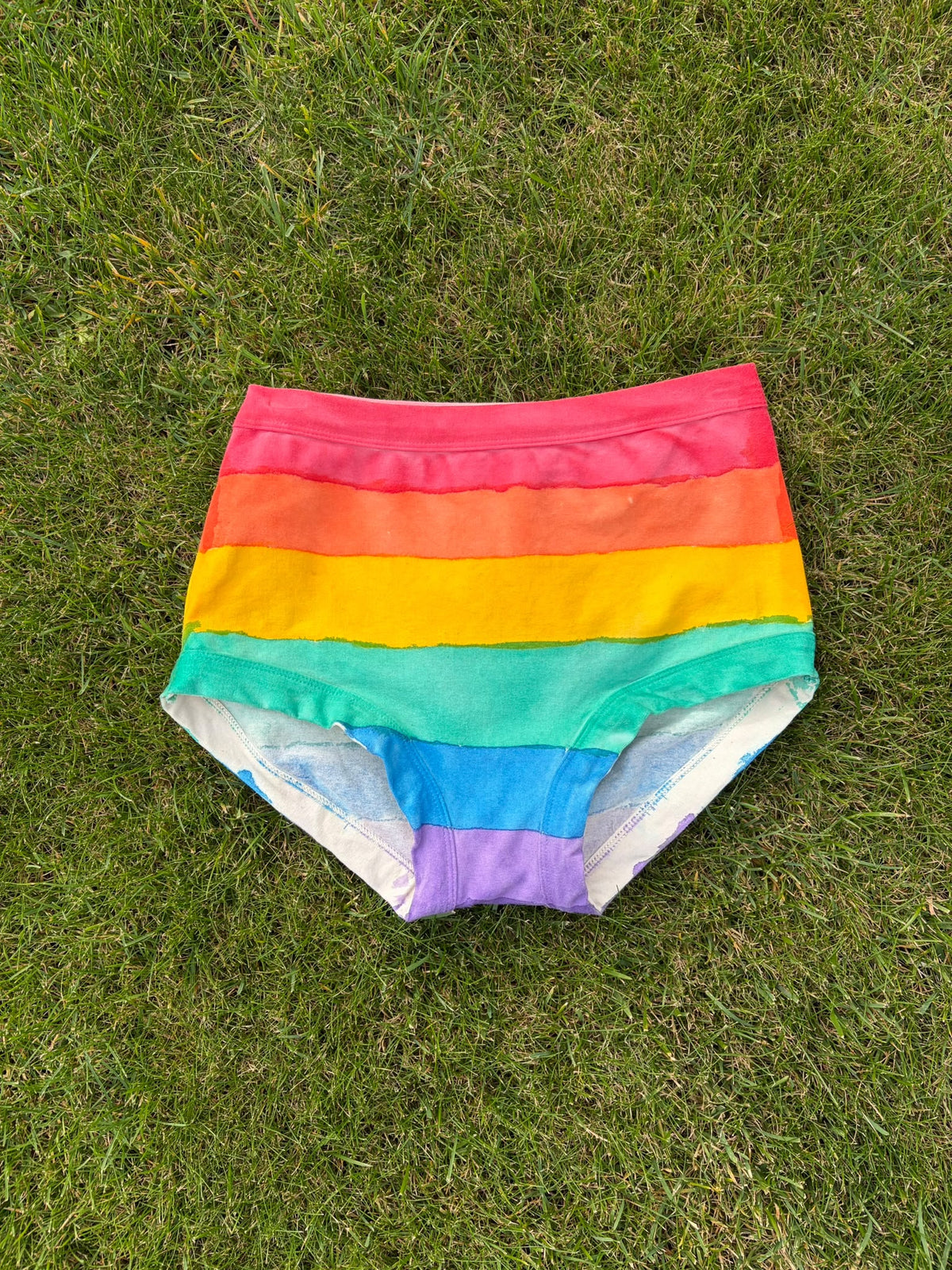 Rainbow-striped original Thunderpants laid flat on green grass, featuring horizontal bands of pink, orange, yellow, green, blue, and purple.