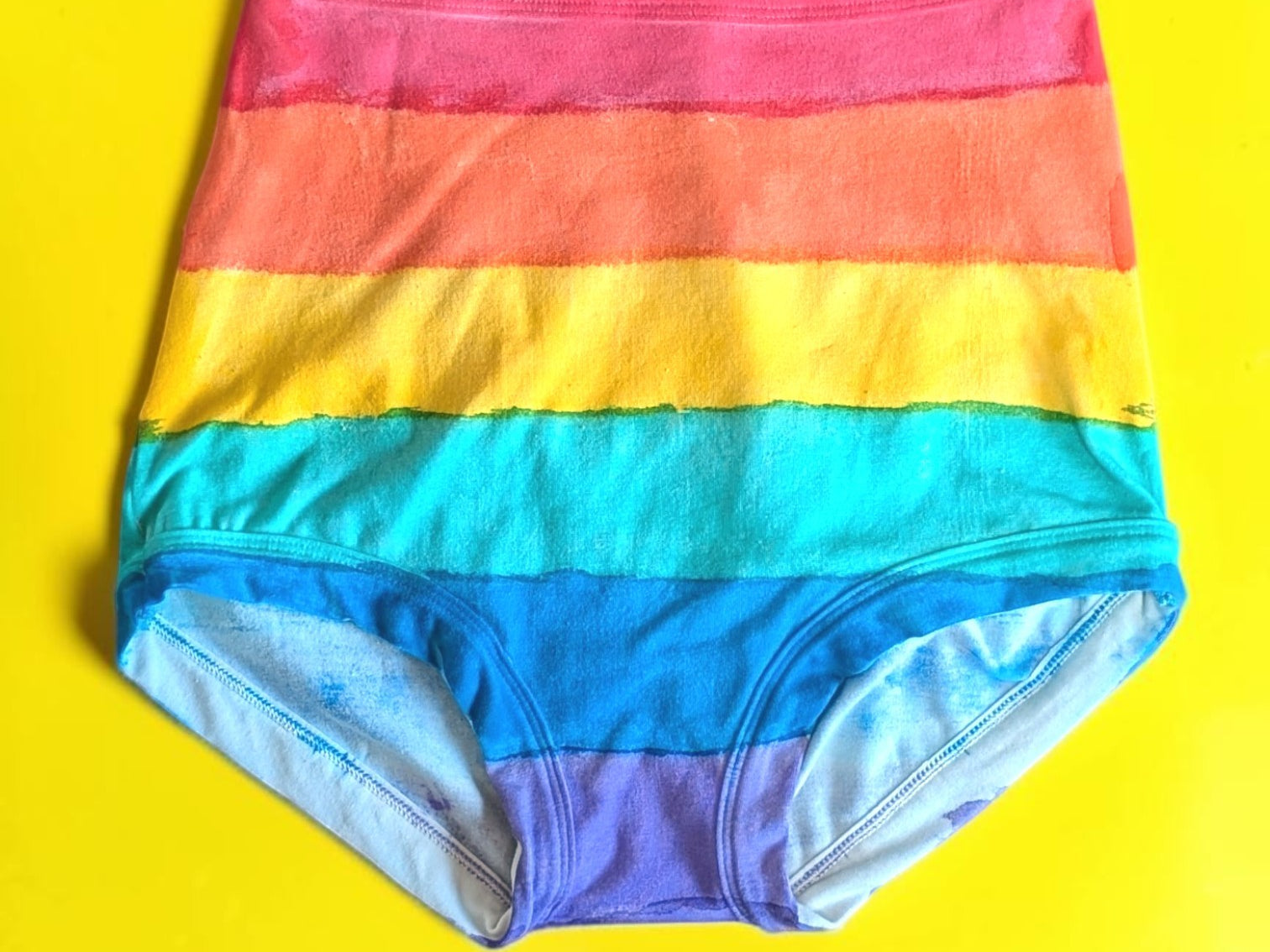 Rainbow-striped high-waisted briefs laid flat on a bright yellow background.