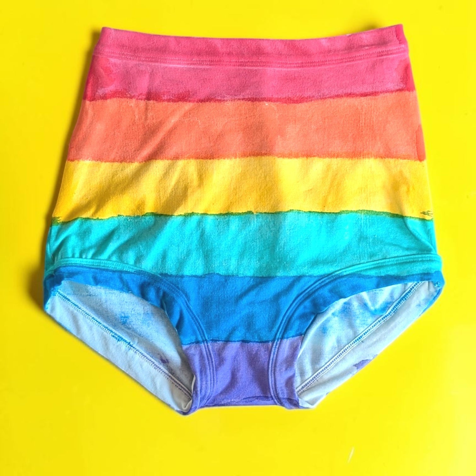 Rainbow-striped high-waisted briefs laid flat on a bright yellow background.