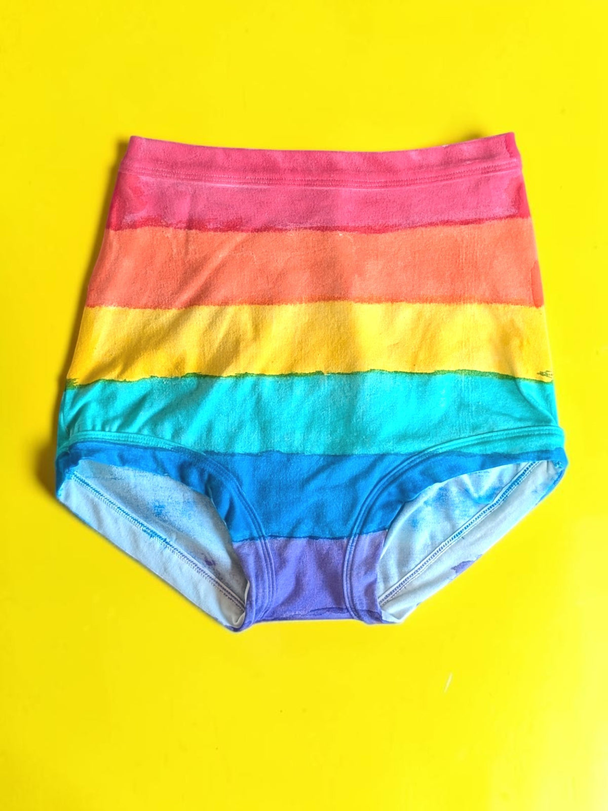 Rainbow-striped high-waisted briefs laid flat on a bright yellow background.