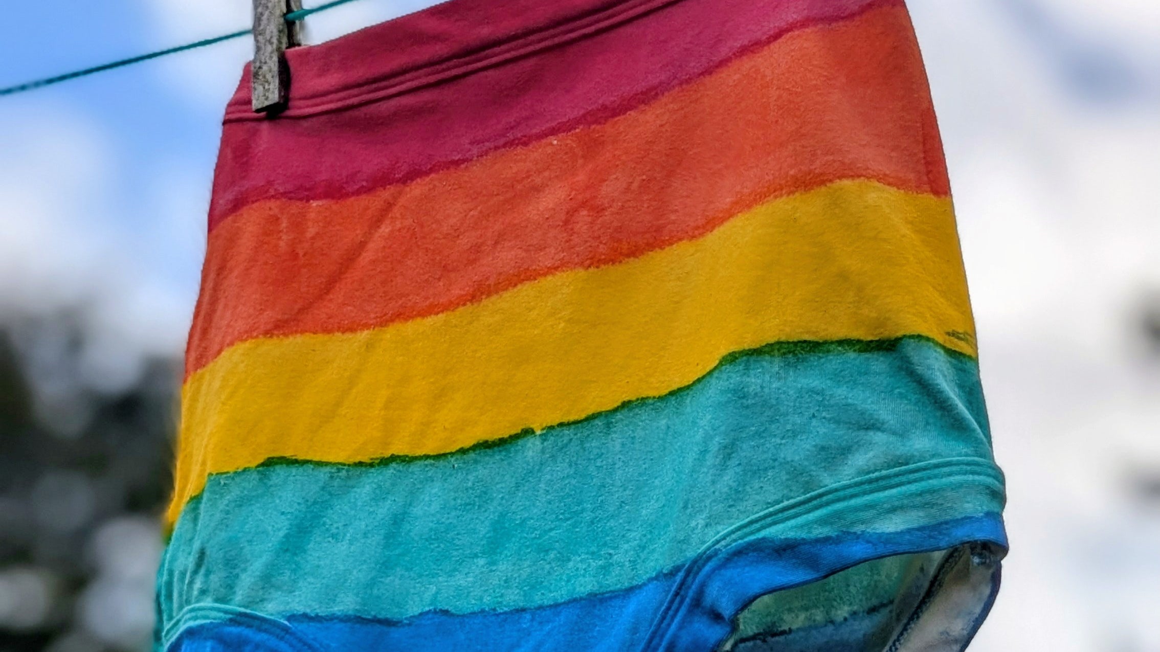 Rainbow-striped underwear hanging from a clothesline outdoors with two wooden clothespins, against a blurred sky and trees background.
