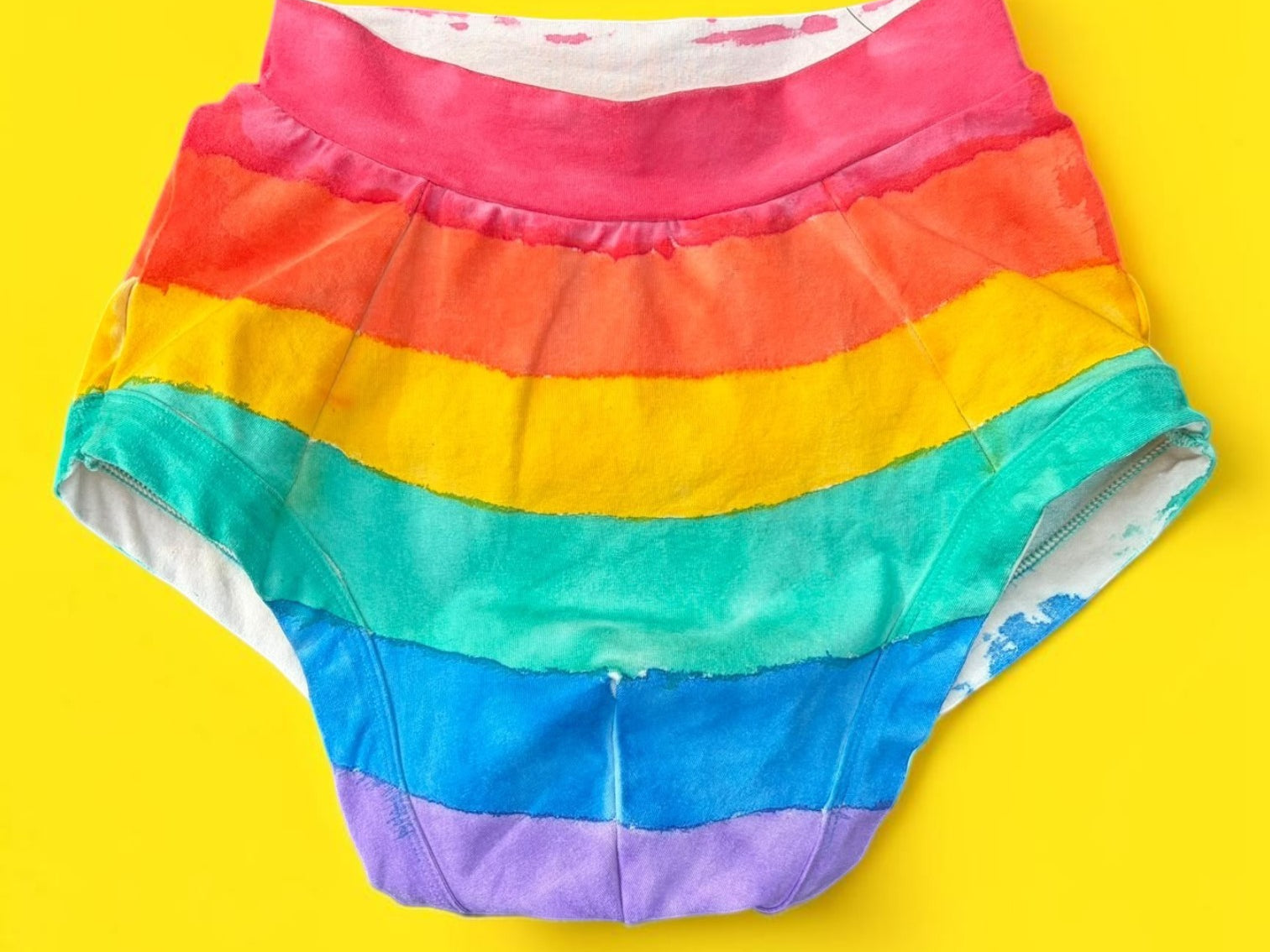 Rainbow-striped briefs with front pouch laid flat on a bright yellow background.