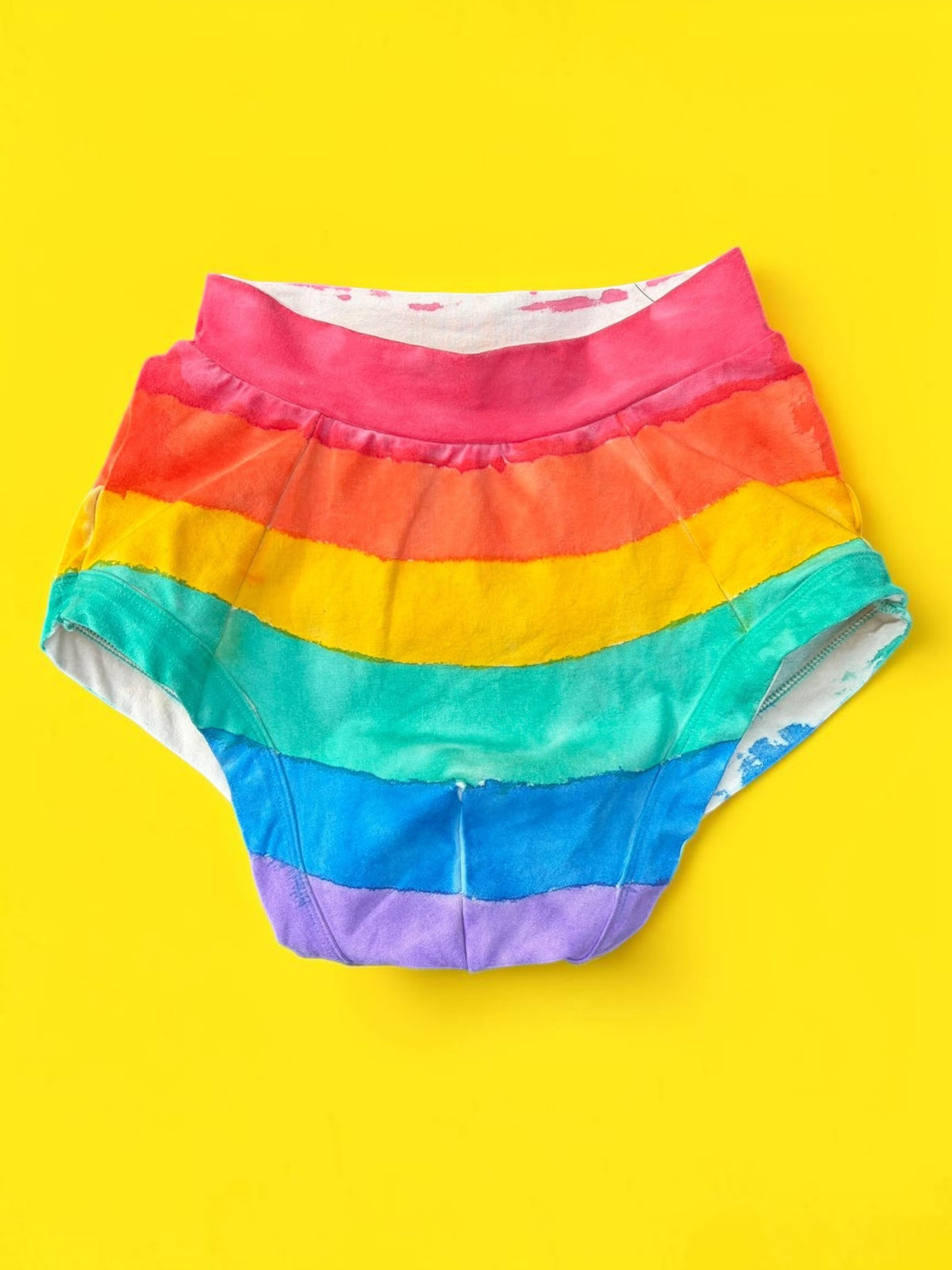 Rainbow-striped briefs with front pouch laid flat on a bright yellow background.