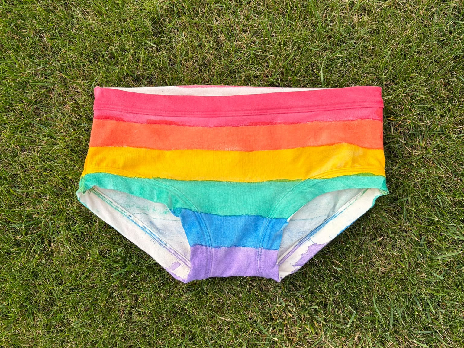 Rainbow striped low rise pants laid flat on a green grass background.