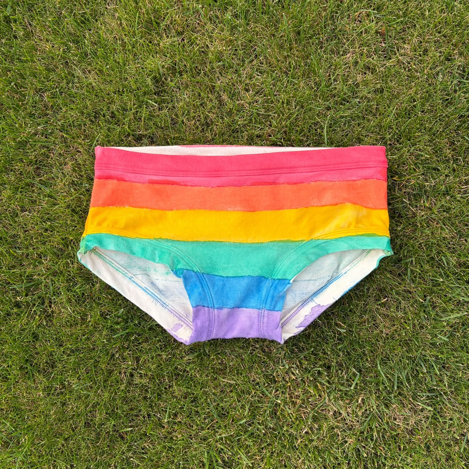 Rainbow striped low rise pants laid flat on a green grass background.