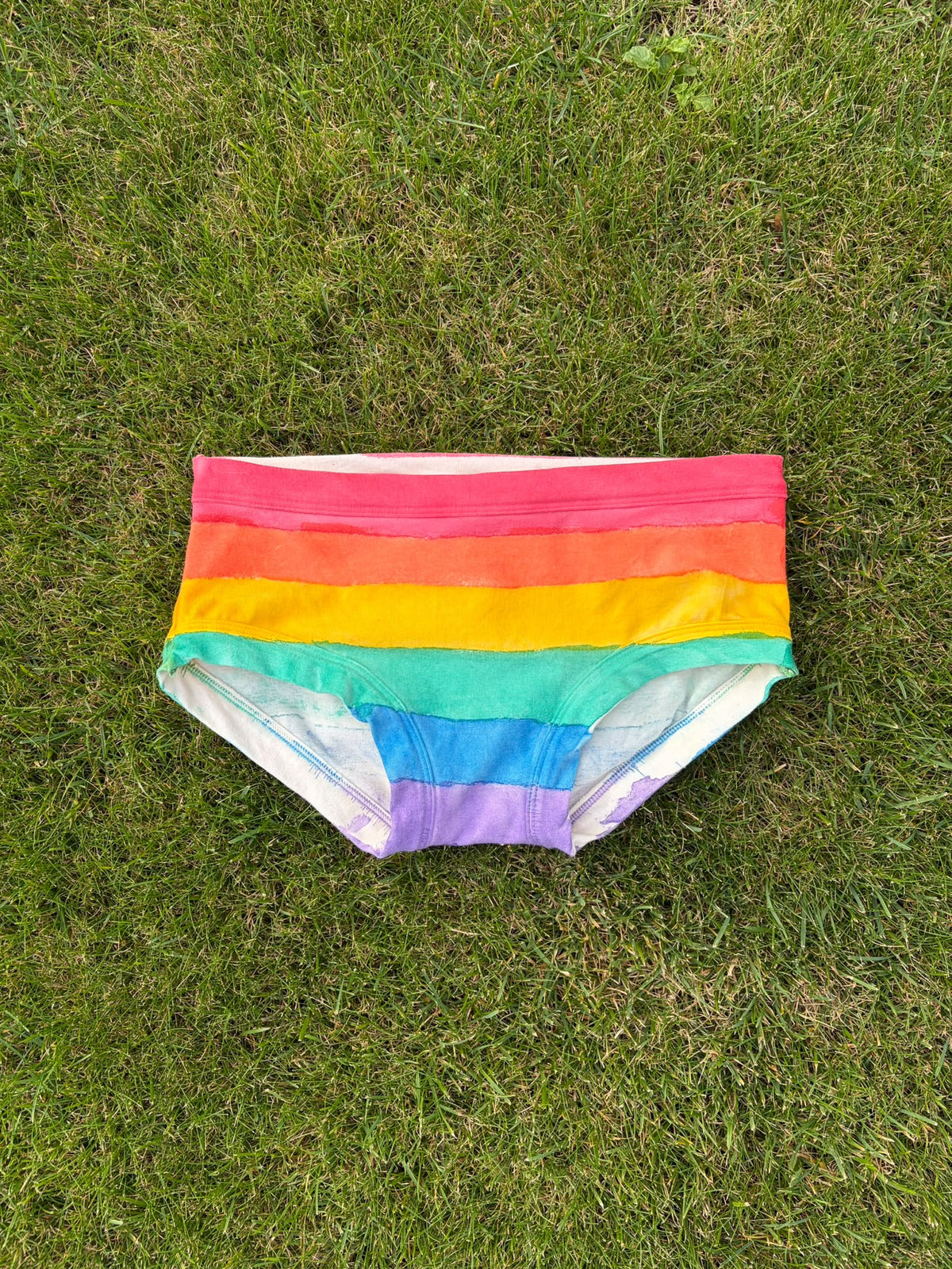 Rainbow striped low rise pants laid flat on a green grass background.
