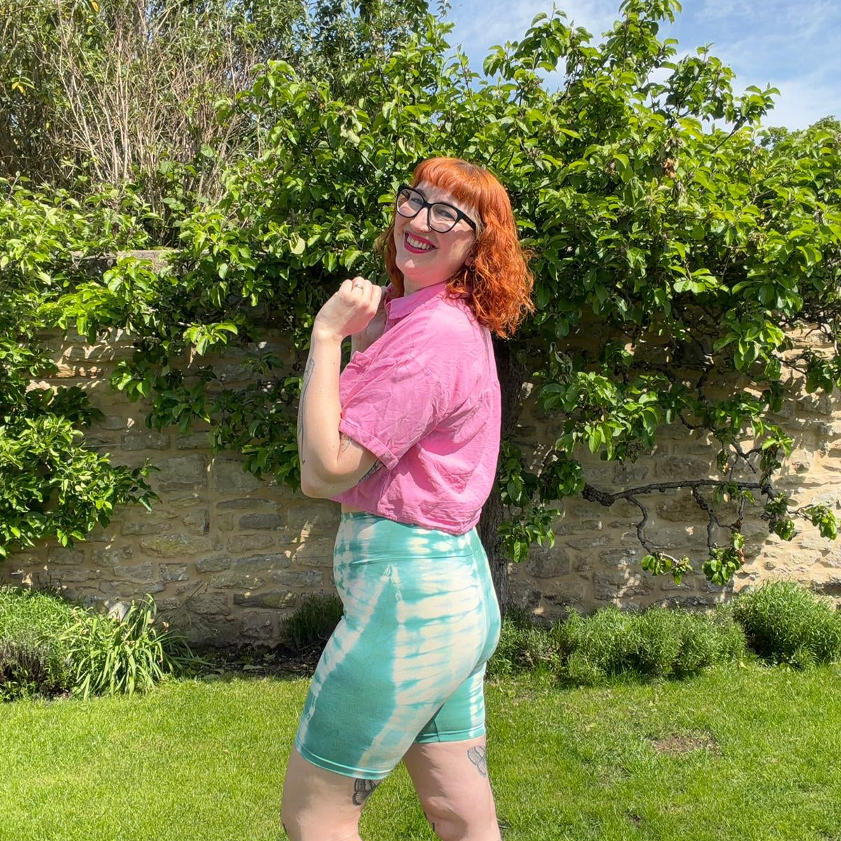 Person wearing a pink shirt and green tie-dye shorts standing outdoors with greenery and a stone wall in the background.