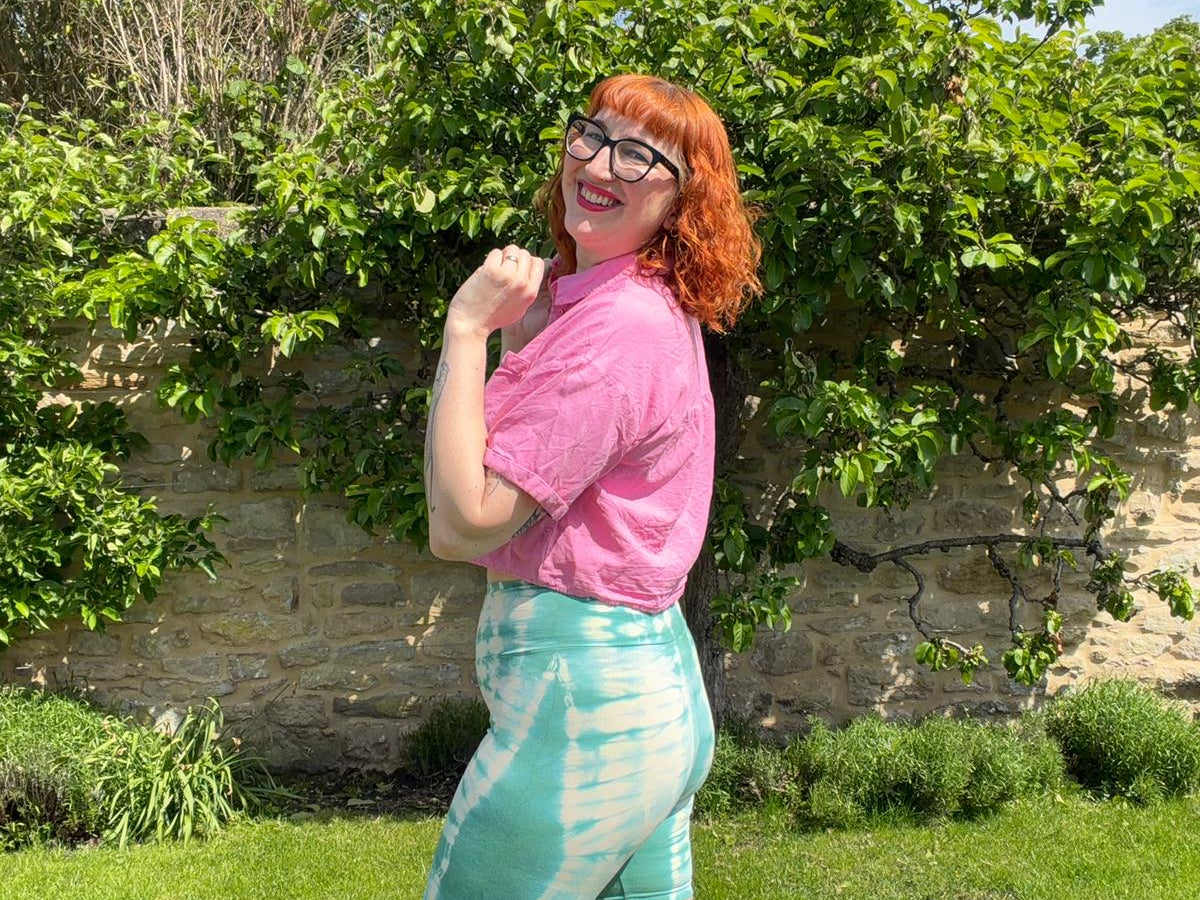 Person wearing a pink shirt and green tie-dye shorts standing outdoors with greenery and a stone wall in the background.