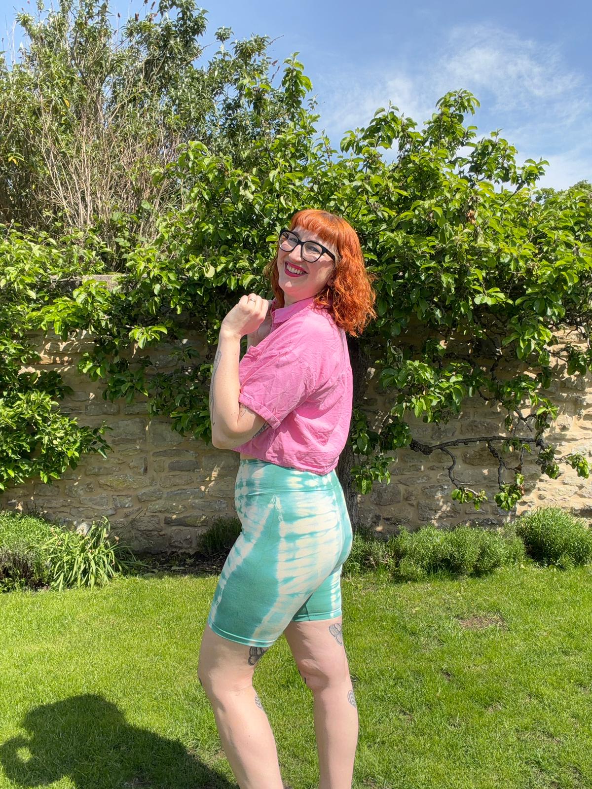 Person wearing a pink shirt and green tie-dye shorts standing outdoors with greenery and a stone wall in the background.